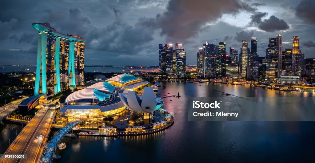 Singapore Urban Cityscape - Skyline at Marina Bay in colorful twilight after sunset. Drone Point of view panorama. Green illuminated Towers. Stitched Panorama DJI Mavic 3 Pro. View over the glowing Singapore Waterfront Modern Skyscrapers and light reflexions under moody twilight skyscape. Singapore, Southeast Asia, Asia
